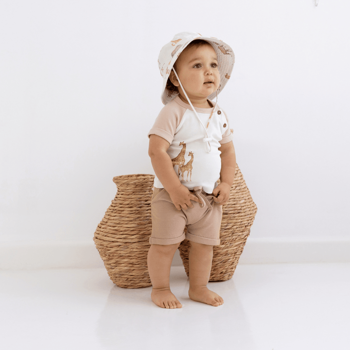 Little boy standing in front of rattan vases wearing a t-shirt with a watercolour giraffe print, taupe sleeves, and coconut buttons, paired with taupe shorts and a bucket hat