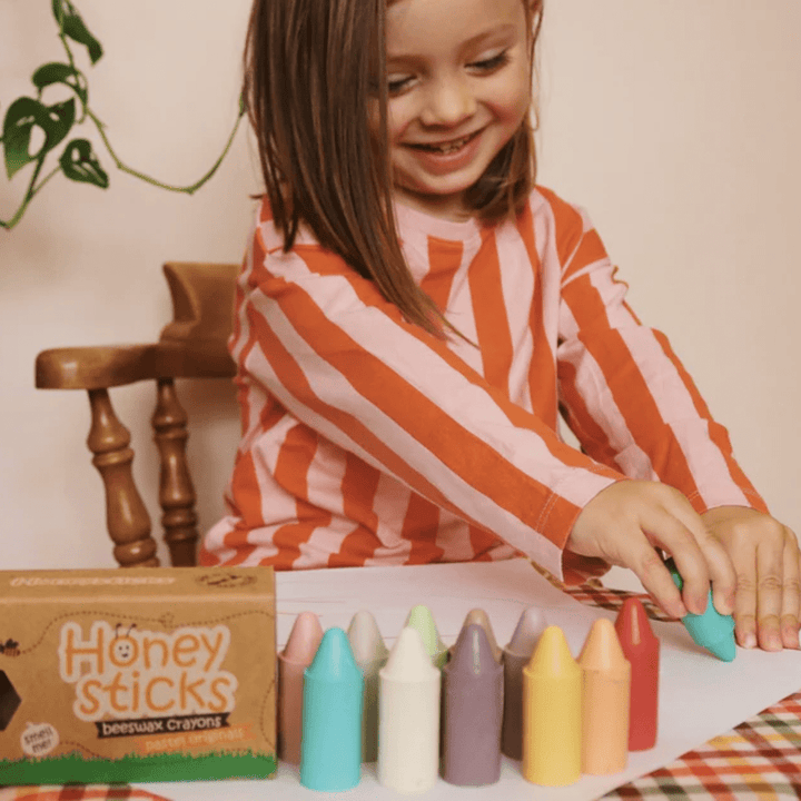 A little girl is playing with Honeysticks Pastel Original Beeswax Crayons on a table.