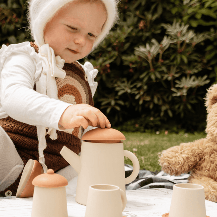 A little girl engaging in imaginative play with a Classical Child Silicone Tea Set on a blanket while using a teddy bear.