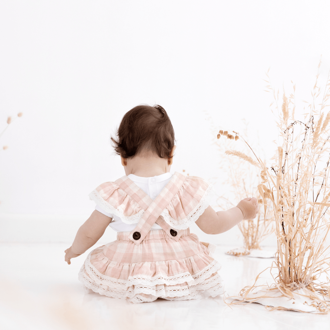 Baby girl sitting down holding on to dried flowers, wearing pink and white gingham playsuit with criss-cross straps, ruffles on the straps, ruffles on the bum, and buttons at the end of the straps
