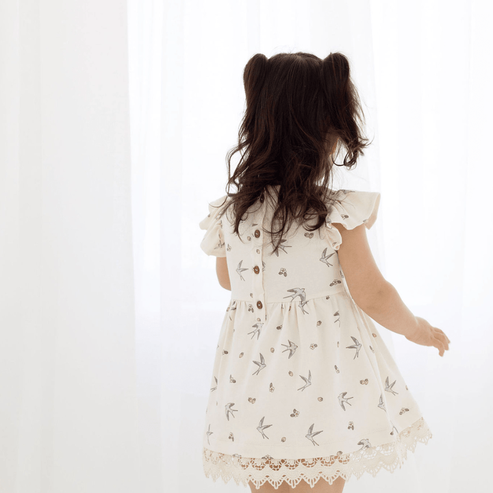 Back view of dark-haired girl with half pigtails wearing an off-white dress with swallow print, with a coconut button opening down the back, short ruffle sleeves, and delicate lace trim
