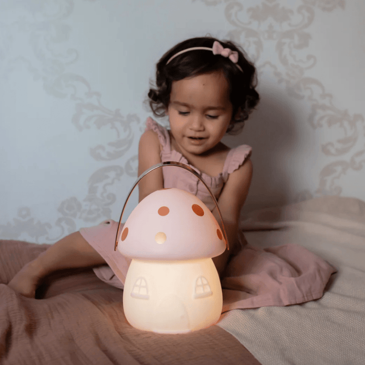 A little girl sitting on a bed with a Little Belle Nightlights Fairy House Carry Lantern in the shape of a pink mushroom, creating a cozy atmosphere resembling a fairy house.