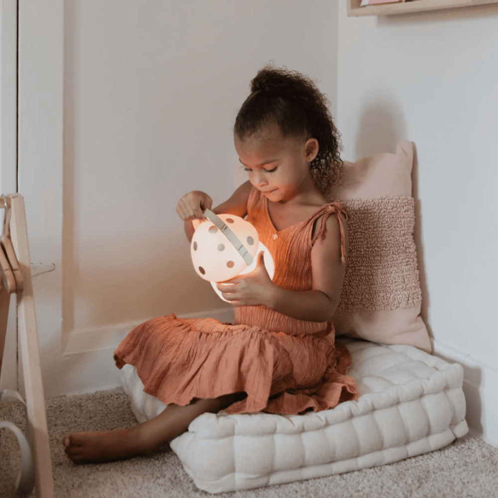 A young child sitting on a cushion and curiously looking inside a lit-up Little Belle Nightlights Mini Fairy House Carry Lantern by Little Belle Nightlights.