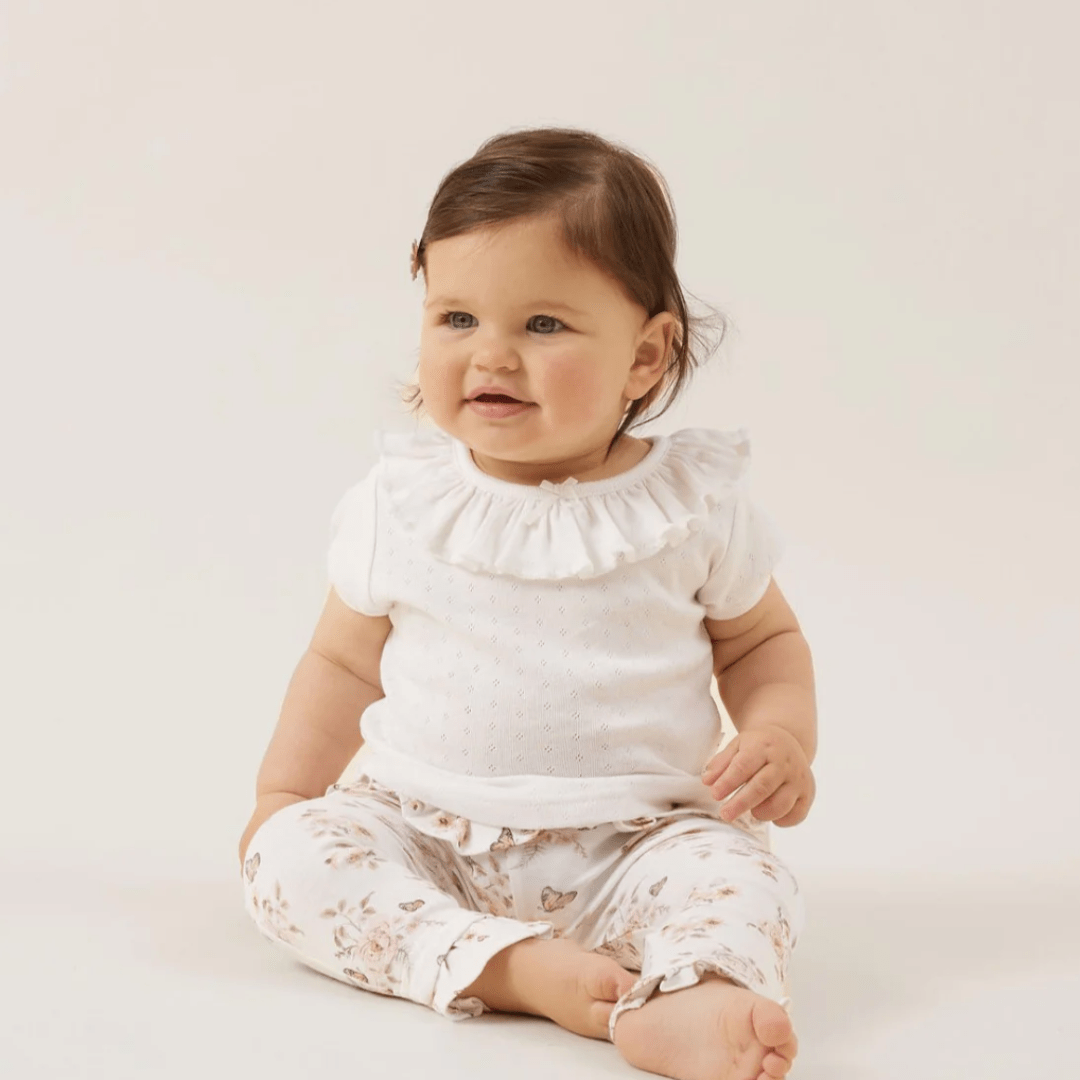 A baby girl is sitting on the floor in an Aster & Oak Organic Cotton Pointelle Ruffle Top.