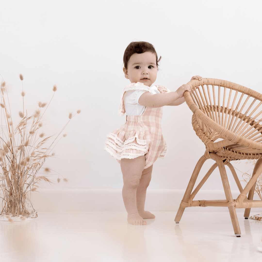 Beautiful baby girl with brown hair and brown eyes looking at camera, wearing pink and white gingham ruffle playsuit, holding on to rattan chair, standing next to dried flowers