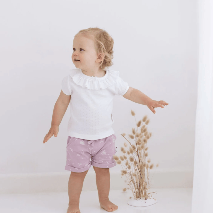 An adorable baby girl wearing Aster & Oak Organic Cotton Harem Shorts, standing in front of a white wall.