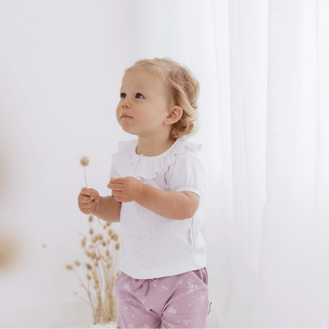 A little girl standing in a white room surrounded by Aster & Oak Organic Cotton Harem Shorts, with an adjustable drawstring waist tie and dandelion flowers.