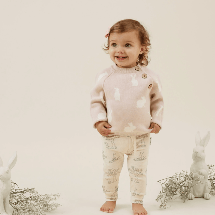 Toddler standing and smiling in Aster & Oak Organic Bunny Luxe Rib Leggings, flanked by rabbit figurines.