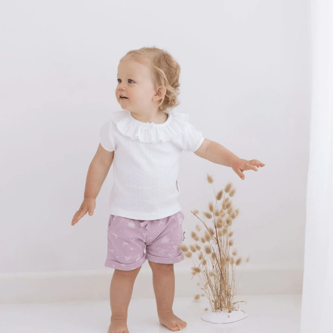 A baby girl wearing an Aster & Oak Organic Cotton Pointelle Ruffle Top and purple shorts.