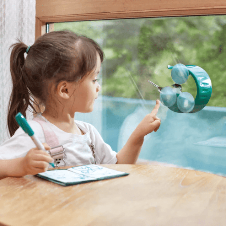 A little girl is sitting at a table and drawing on a window while birdwatching with the Hape Bird Feeder.