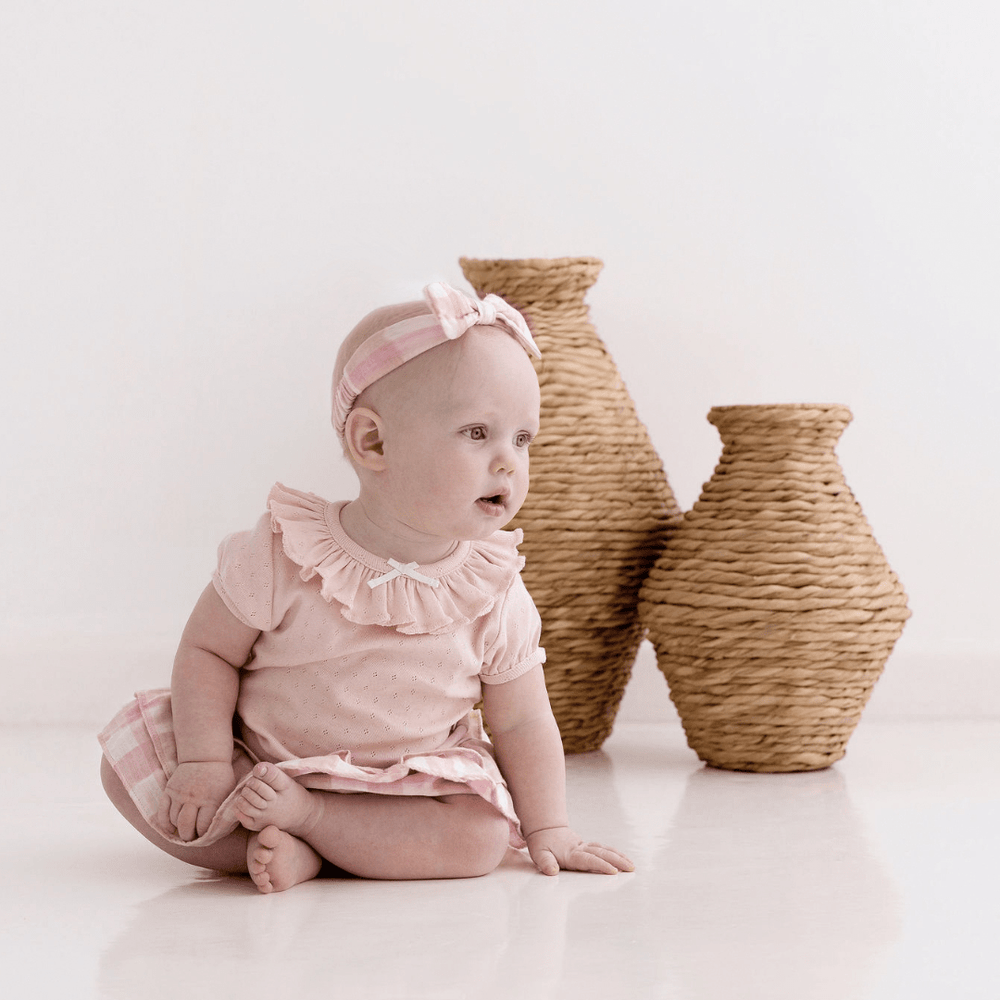 Baby Girl Sitting Cross-Legged In Front Of Rattan Vases, Wearing Pink & White Gingham Baby Headband With Bow
