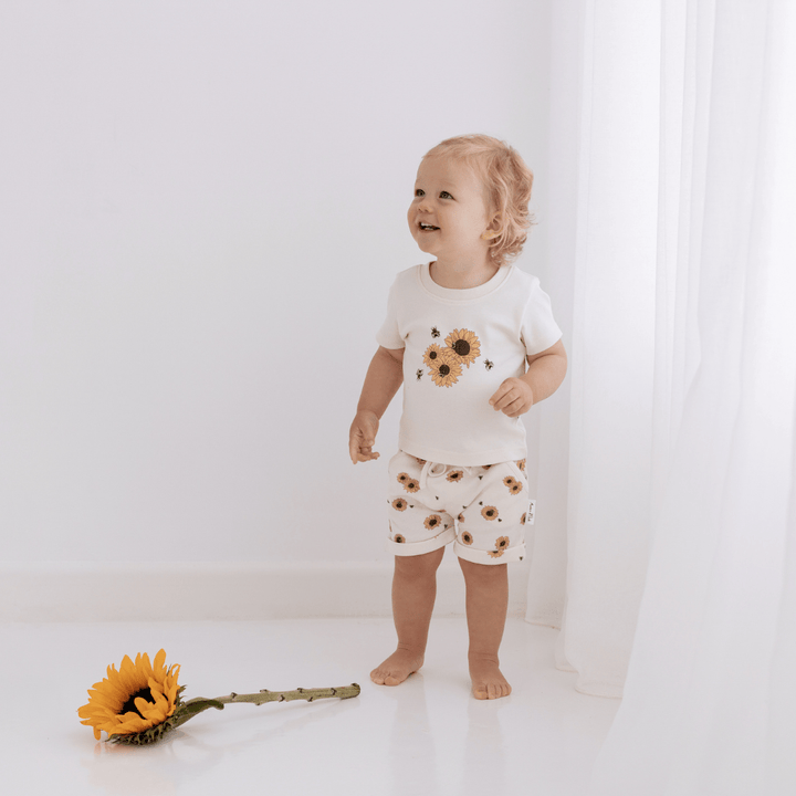 A baby standing next to a sunflower in a white room wearing comfortable Aster & Oak Organic Cotton Harem Shorts made from organic cotton.