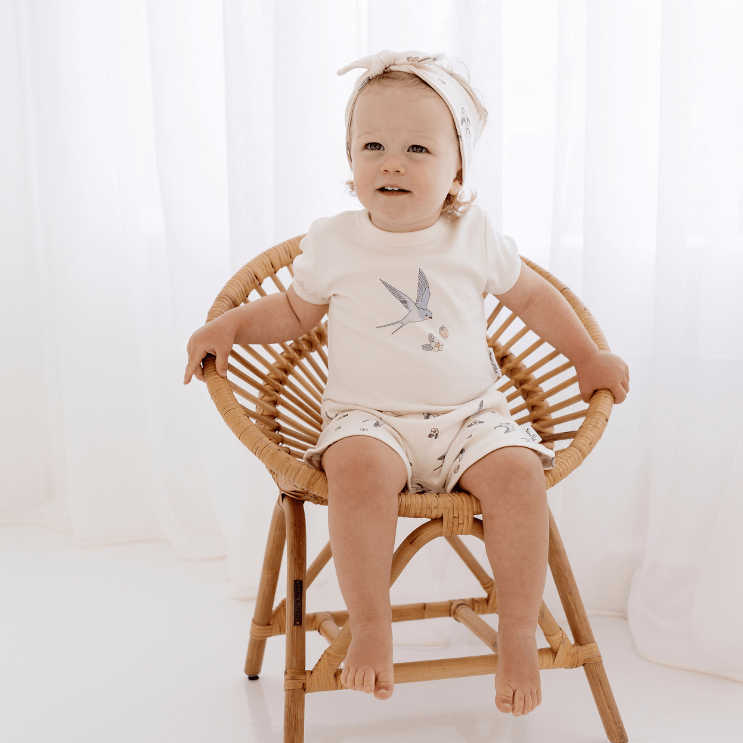 A baby sitting in a wicker chair wearing Aster & Oak Organic Cotton Harem Shorts with an adjustable drawstring waist tie.