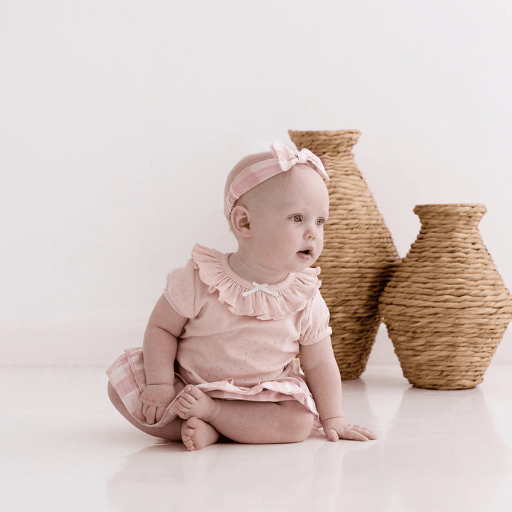 An Aster & Oak Organic Cotton Pointelle Ruffle Top baby sitting on the floor next to a wicker basket.