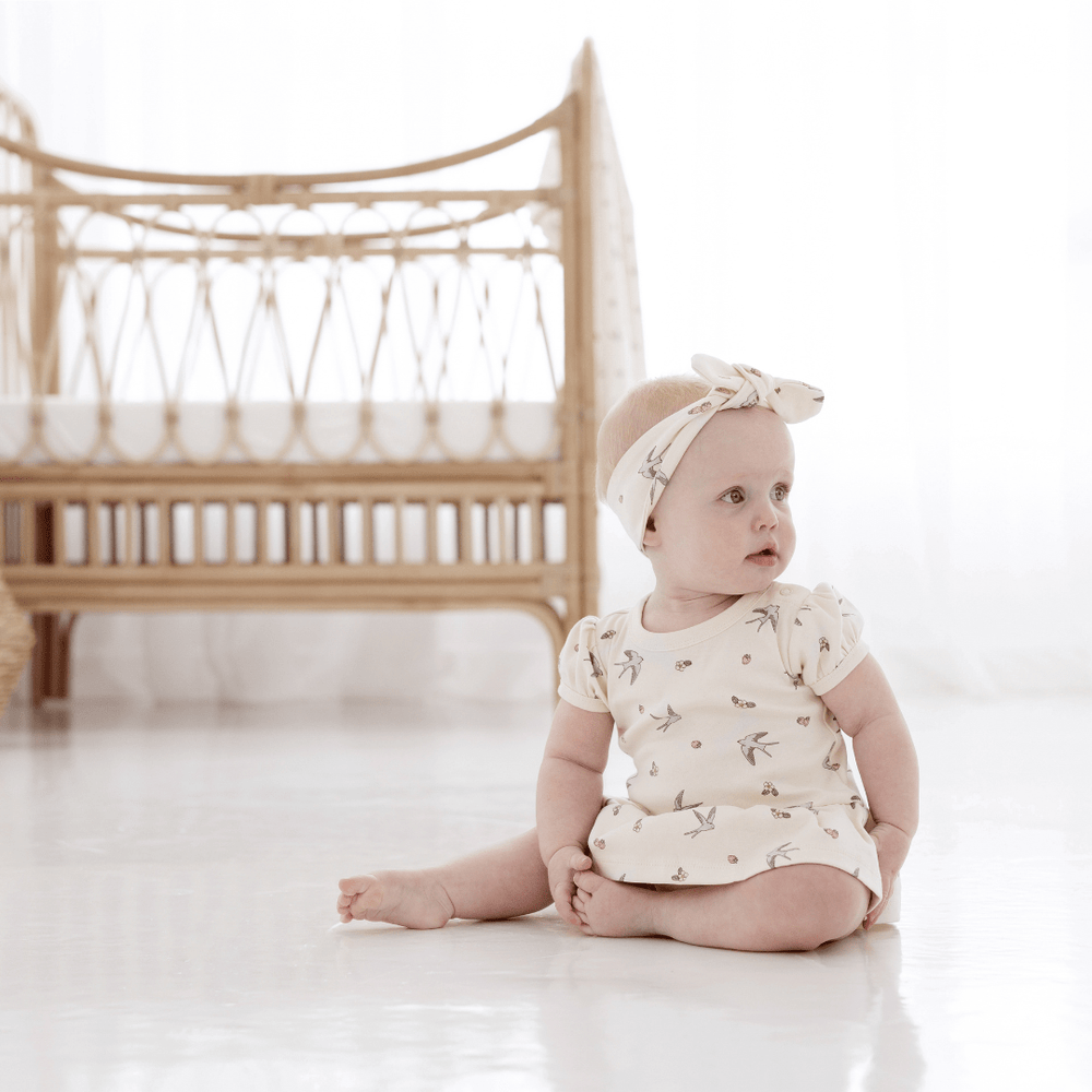 Baby looking over her left shoulder, sitting in front of a rattan cot, wearing a natural-coloured baby onesie with built-in skirt, and a matching headband. Both have a delicate hand-illustrated all-over swallow and strawberry print on them.