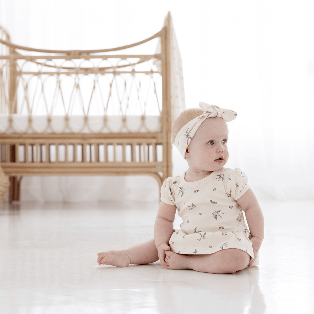 Baby looking over her left shoulder, sitting in front of a rattan cot, wearing a natural-coloured baby onesie with built-in skirt, and a matching headband. Both have a delicate hand-illustrated all-over swallow and strawberry print on them.