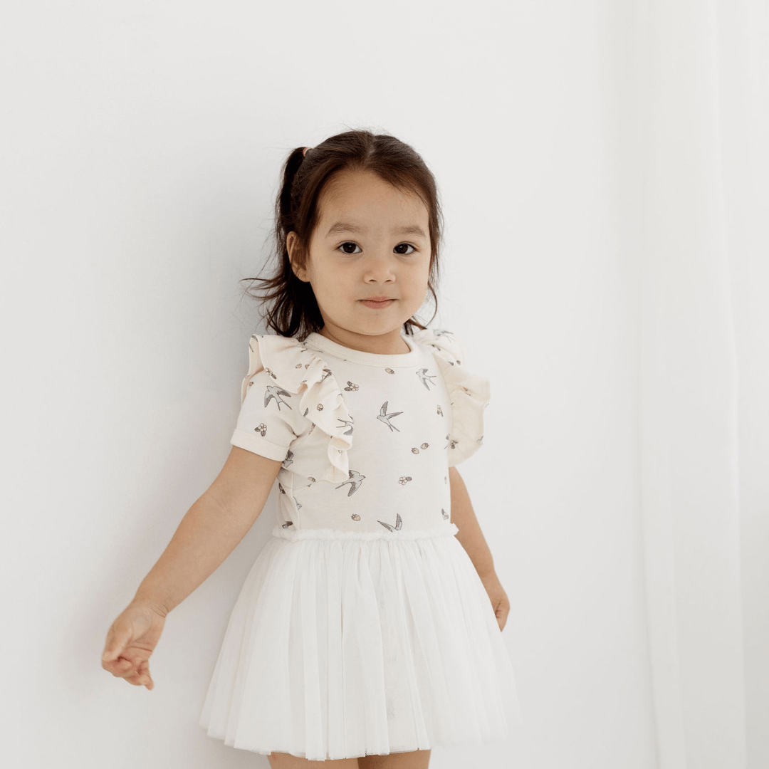 Brown-haired little girl standing in front of a sheer white curtain, wearing a beautiful dress for babies and kids featuring a delicate hand-drawn swallow and strawberry print on the top half, short sleeves with a ruffle at the shoulder, and a tulle skirt