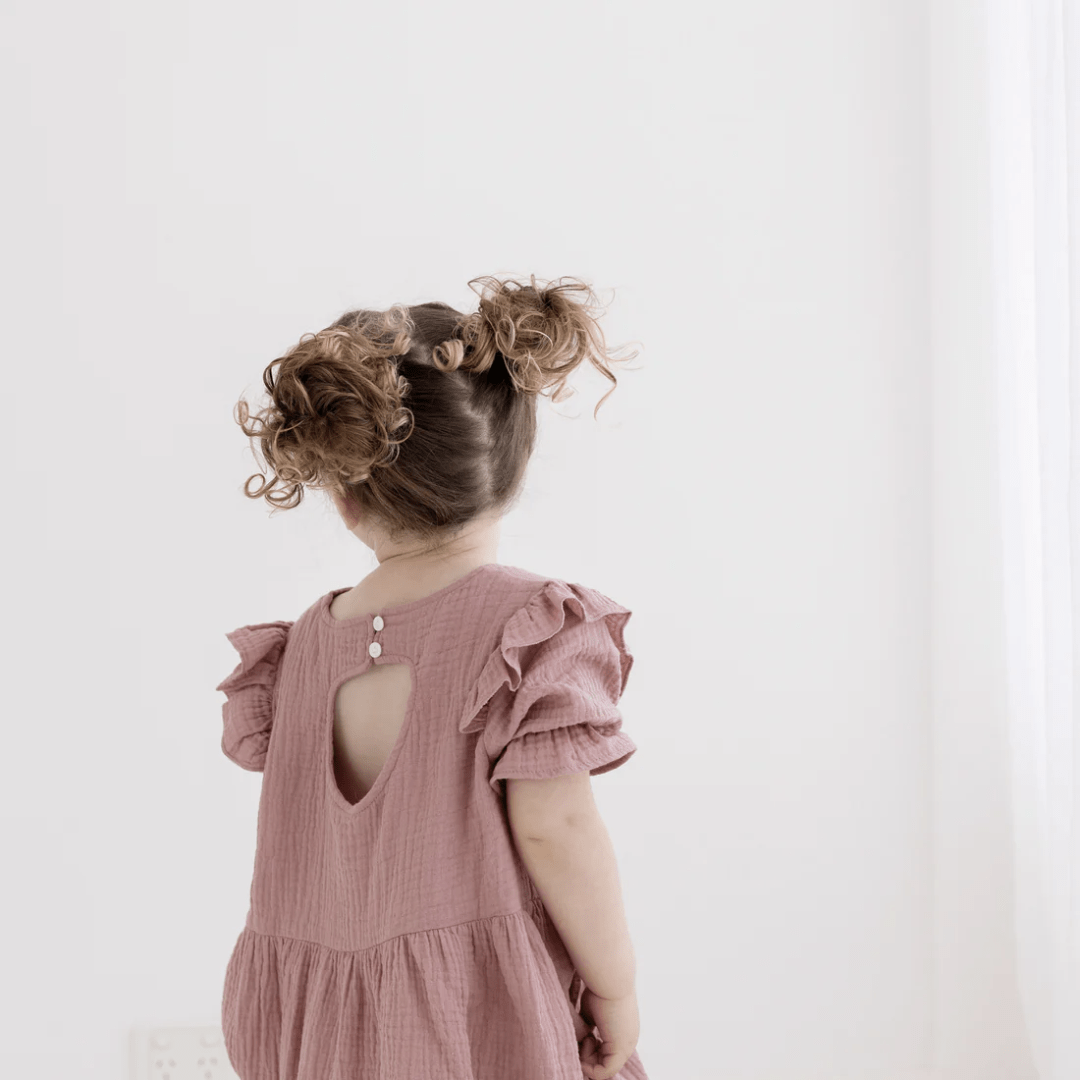A little girl in an Aster & Oak Organic Rosette Muslin Ruffle Dress - LUCKY LASTS - 4 YEARS & 5 YEARS ONLY standing in a white room.