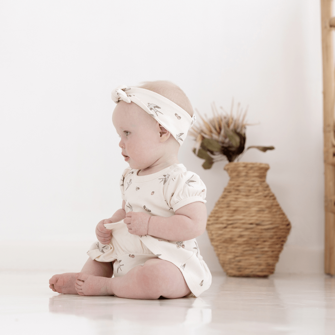 Baby girl sitting on floor holding on to the skirt of her skirt onesie. The onesie is a natural colour, with a delicate swallow and strawberry print. She has a matching headband with a bow and is sitting in front of a rattan floor vase with a dried flower in it.