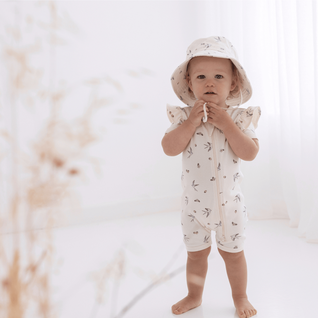 A baby wearing an Aster & Oak Organic Cotton Flutter Sleeve Zip Romper and hat in a white room.