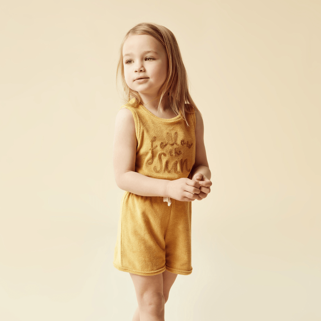 A little girl in a Wilson & Frenchy Organic Terry Kids Playsuit (Multiple Variants) from Wilson & Frenchy standing on a beige background.
