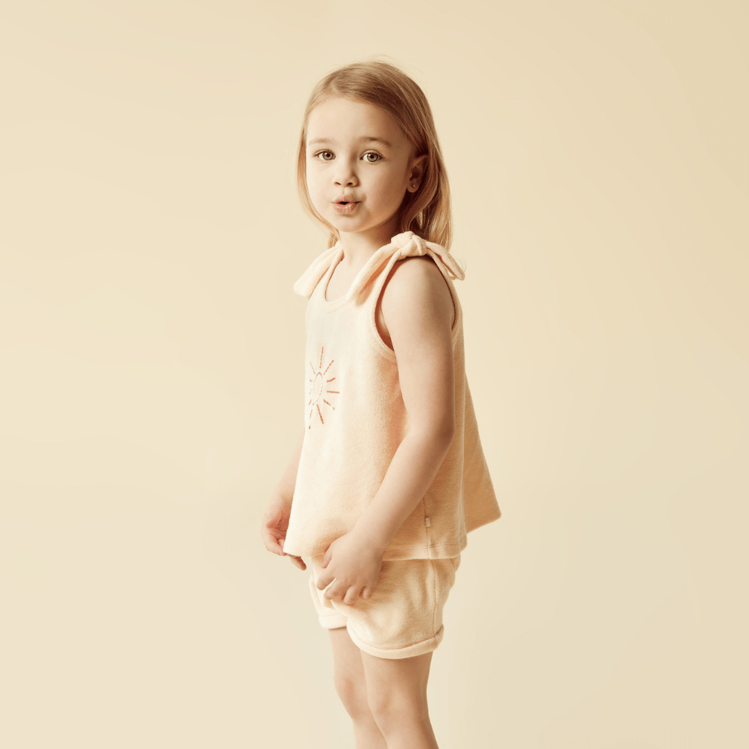 A little girl wearing a peach-colored Wilson & Frenchy Organic Terry Tie Kids Singlet made of organic cotton with tie-ups on her shorts.