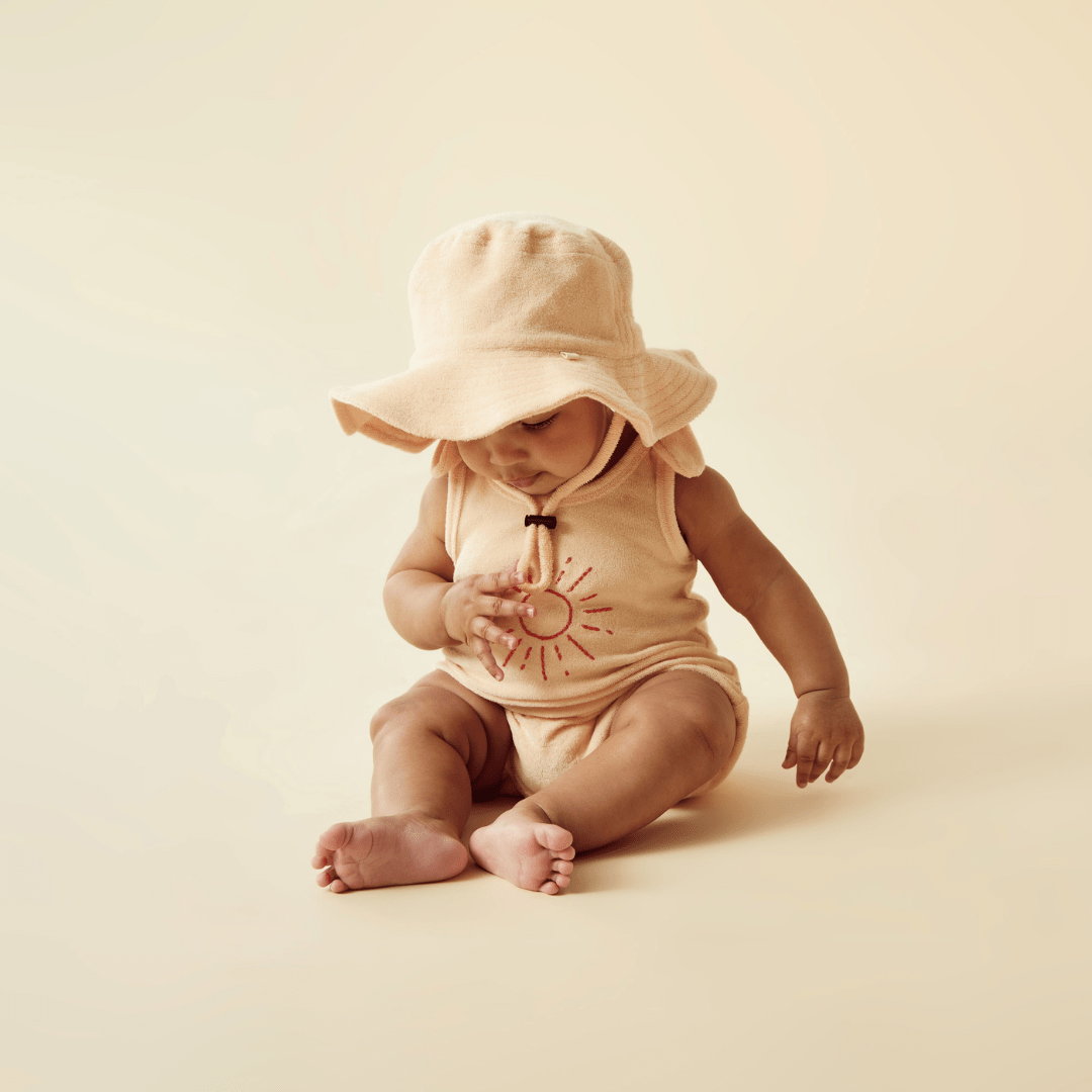 A baby in a hat, wearing Wilson & Frenchy's Wilson & Frenchy Organic Terry Tie Singlet, sitting on the ground. The singlet is made from GOTS-certified organic fabric.