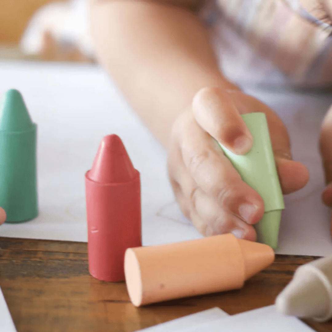 A child is using Honeysticks Pastel Original Beeswax Crayons from the brand Honeysticks to create vibrant and long-lasting drawings on a table. These environmentally sustainable crayons offer rich, lasting colors while being an eco-friendly choice.