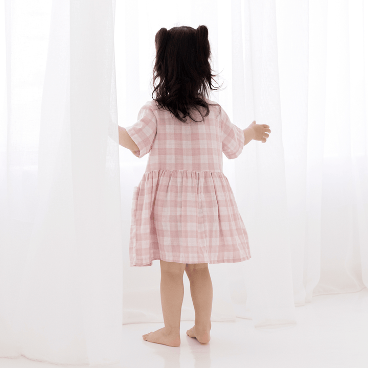 Dark-Haired Girl Standing Looking Out Window With A White Sheer Curtain, Wearing Pink & White Gingham Dress