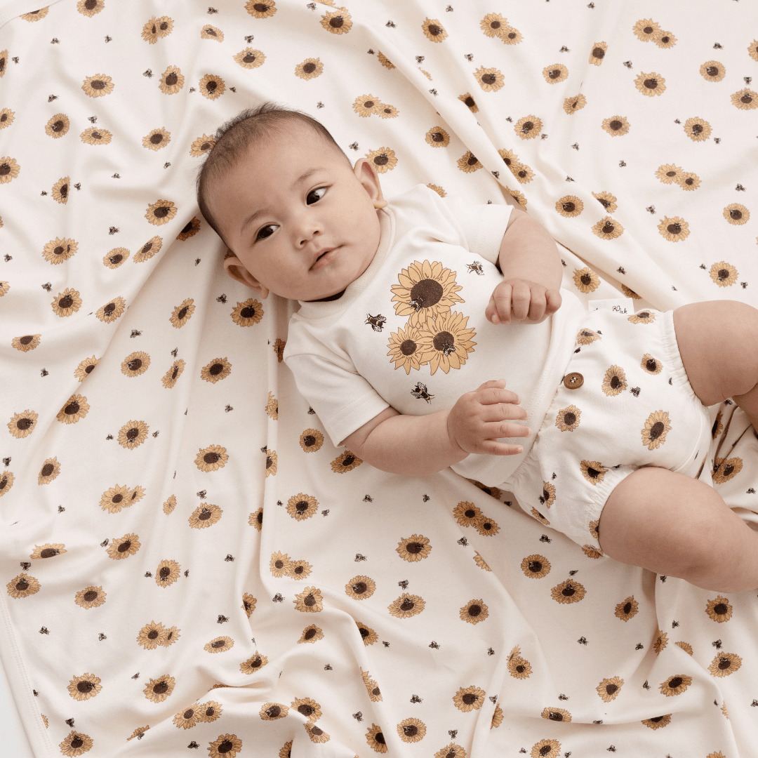 A baby swaddled in an Aster & Oak Organic Cotton Baby Swaddle Wrap featuring a hand-illustrated sunflower print.