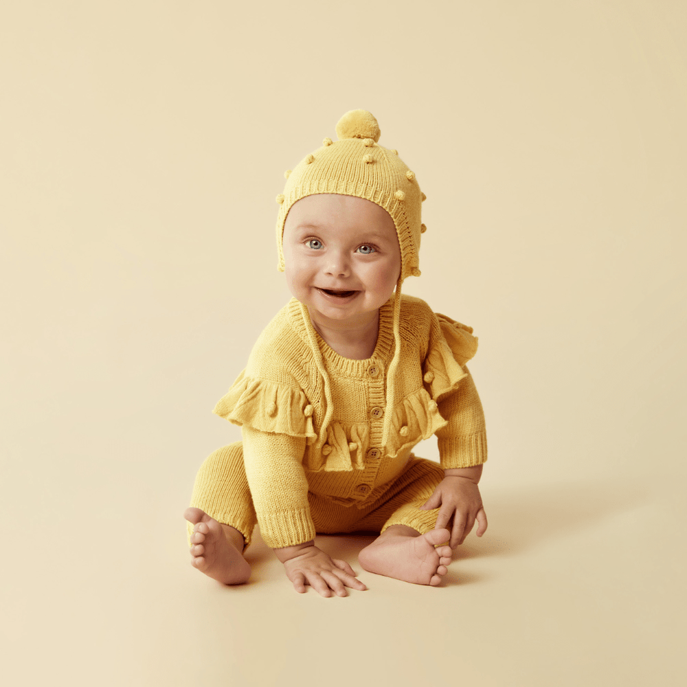 A smiling baby dressed in a yellow knitted outfit with a Wilson & Frenchy Knitted Bauble Bonnet sits against a beige background.