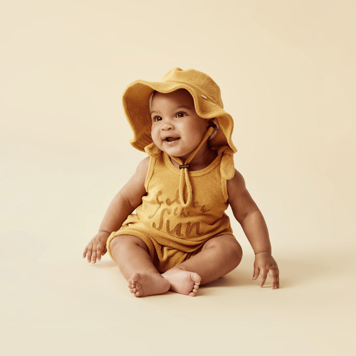 A baby wearing a yellow hat and romper made from Wilson & Frenchy Organic Terry Tie Singlet (Multiple Variants) made with GOTS-certified organic fabric.