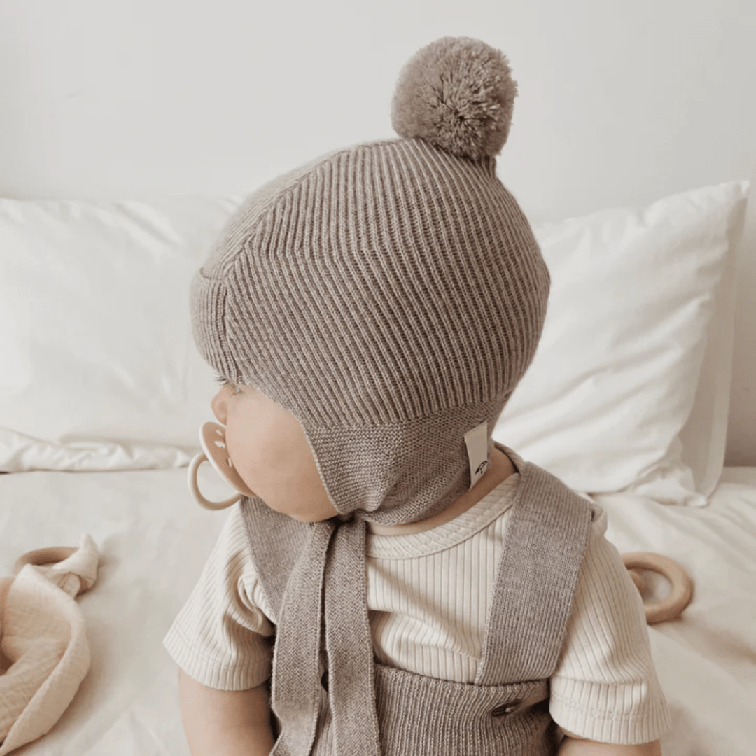 A baby sitting on a bed wearing a Saga Copenhagen Merino Bonnet with a pom pom.
