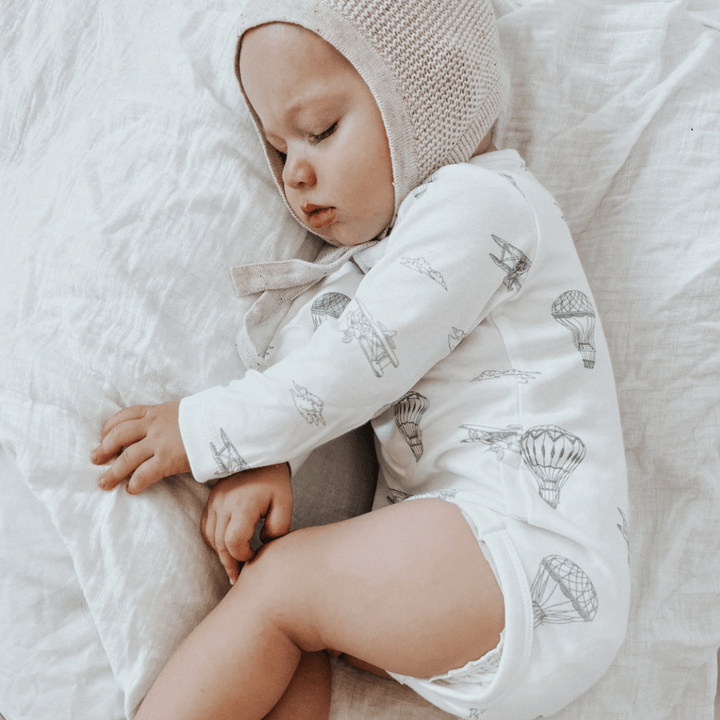 A baby wearing a beanie and an Aster & Oak Organic Henley Long-Sleeved Onesie with a hot air balloon print is sleeping peacefully on a light-colored fabric.