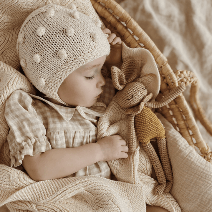 An eco-friendly baby peacefully sleeping in a basket, accompanied by a Patti Oslo Organic Cotton Bianca Bunny stuffed toy made from organic cotton.