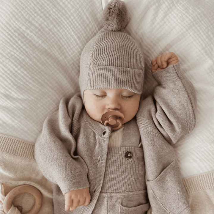 A baby peacefully sleeping on a bed with a wooden toy, wearing a cozy Saga Copenhagen Merino Bonnet by Saga Copenhagen.