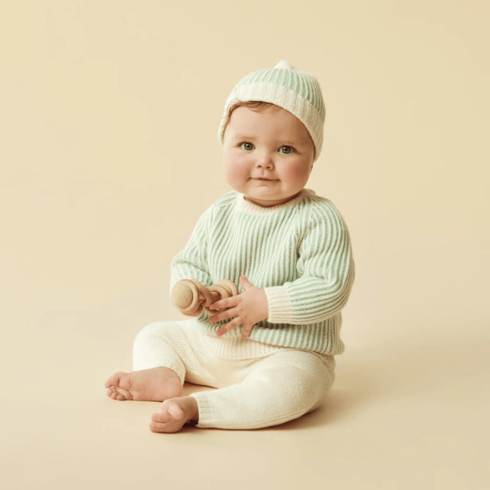 A baby wearing a green and white striped outfit with a matching Wilson & Frenchy Knitted Ribbed Hat, sitting and holding a wooden toy.