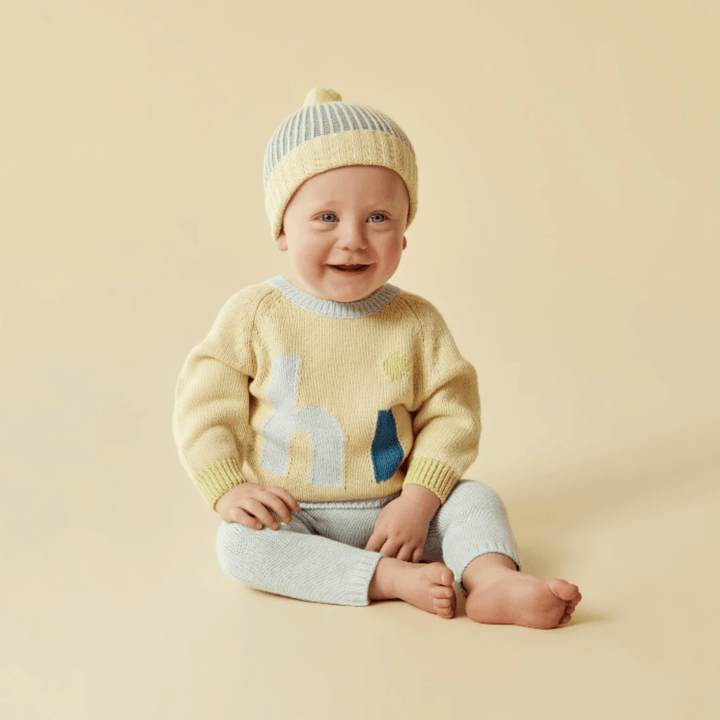 A smiling baby wearing a striped beanie and a yellow Wilson & Frenchy knitted jacquard jumper with a giraffe design sits against a beige background.
