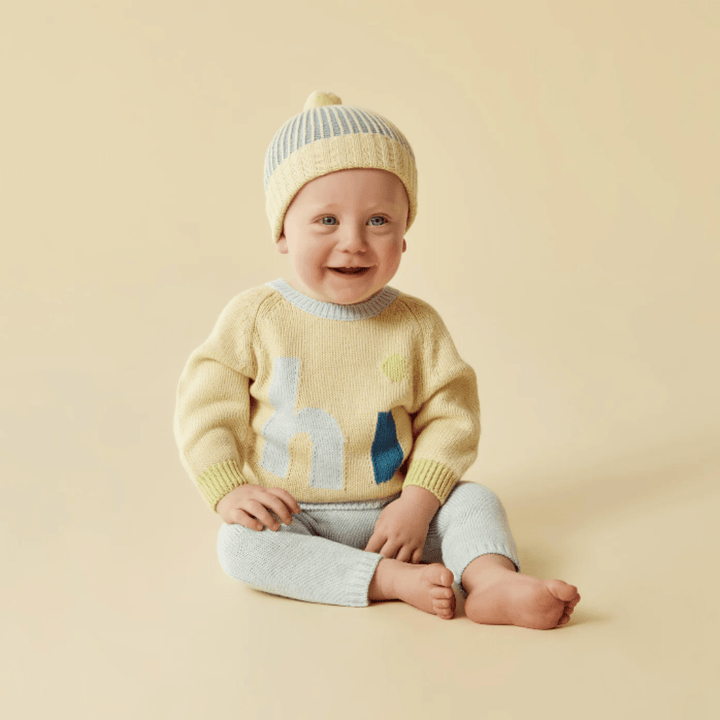 A smiling baby wearing a Wilson & Frenchy Knitted Ribbed Hat and a yellow sweater with a blue giraffe pattern, seated against a beige background.