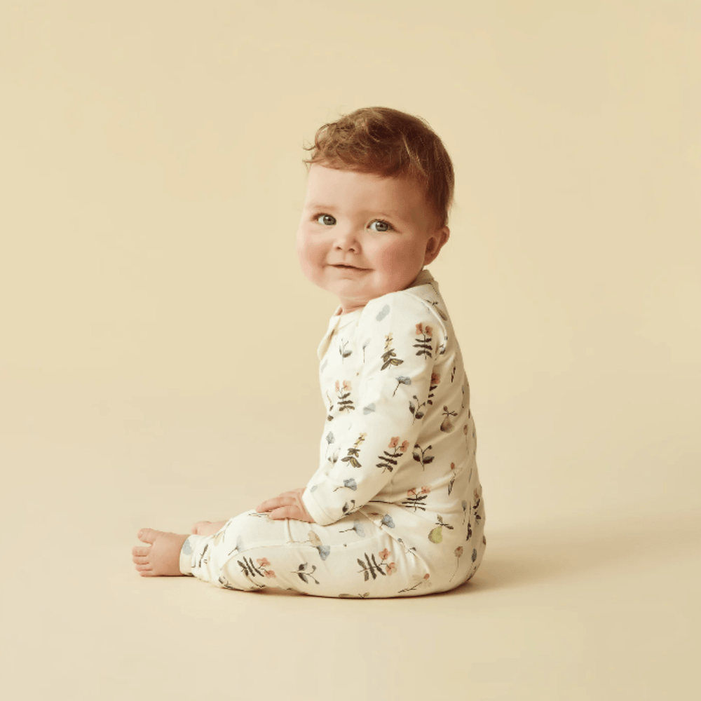 A smiling newborn sitting on a beige background, wearing Wilson & Frenchy Organic Baby Pyjamas with a hand-illustrated print.