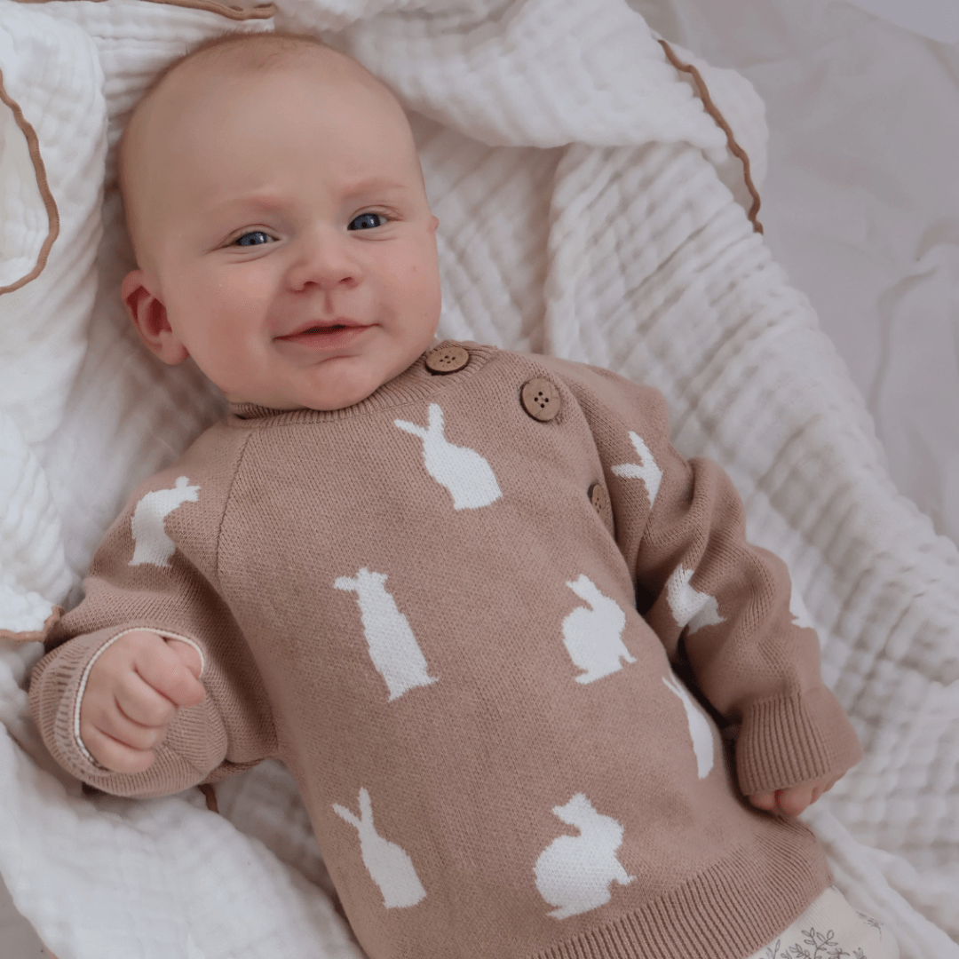 A smiling baby lying down wearing an Aster & Oak Organic Knit Bunny Jumper with white rabbit patterns.
