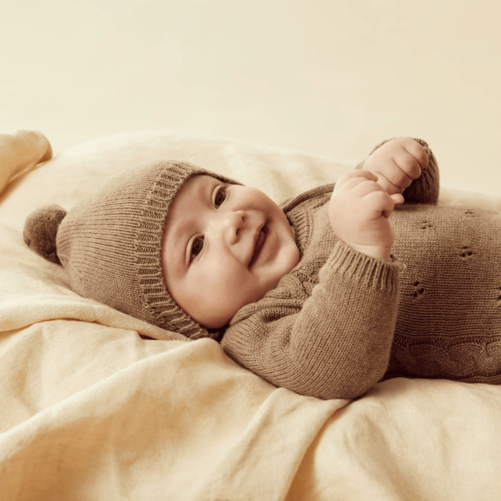A smiling baby wearing a Wilson & Frenchy Knitted Bonnet and onesie.