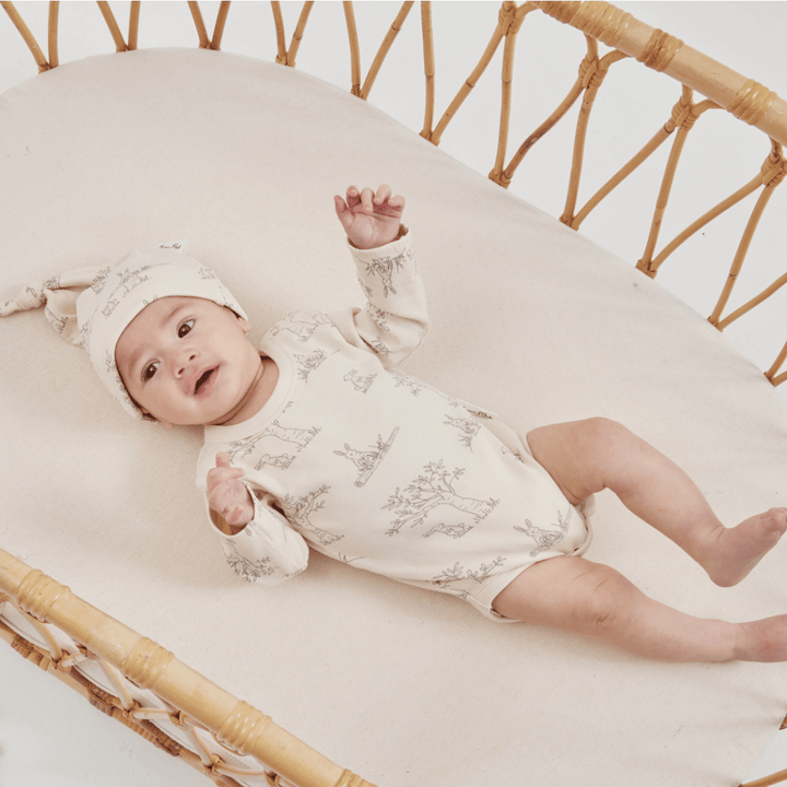 Infant lying in a wicker bassinet wearing an Aster & Oak Organic Bunny Luxe Rib Long-Sleeved Onesie and matching hat.
