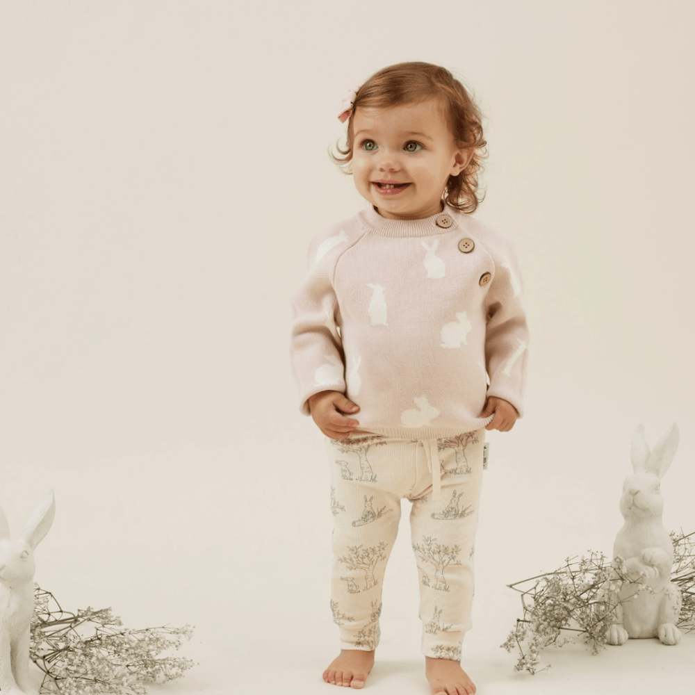 A toddler with a pink bow in her hair, wearing an Aster & Oak Organic Knit Bunny Jumper and patterned pants, stands smiling in a studio setting with rabbit figurines around.