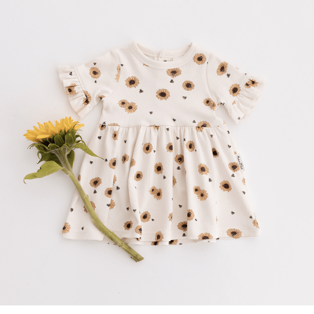 Dress for babies and kids featuring yellow sunflowers on an off-white background, ruffle sleeves, and a full skirt, featured with a bright yellow sunflower lying to the side