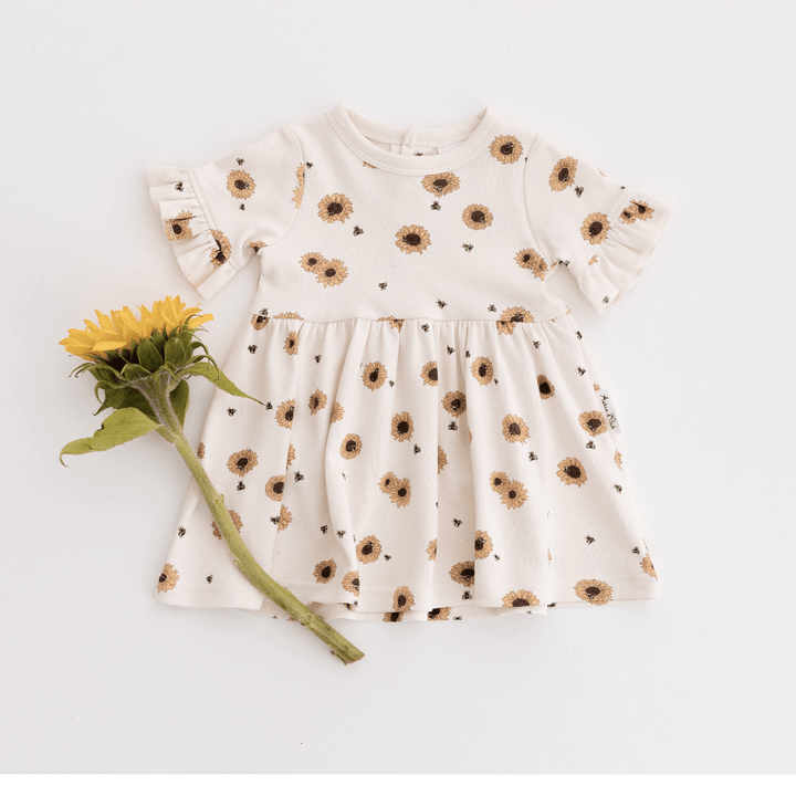 Dress for babies and kids featuring yellow sunflowers on an off-white background, ruffle sleeves, and a full skirt, featured with a bright yellow sunflower lying to the side