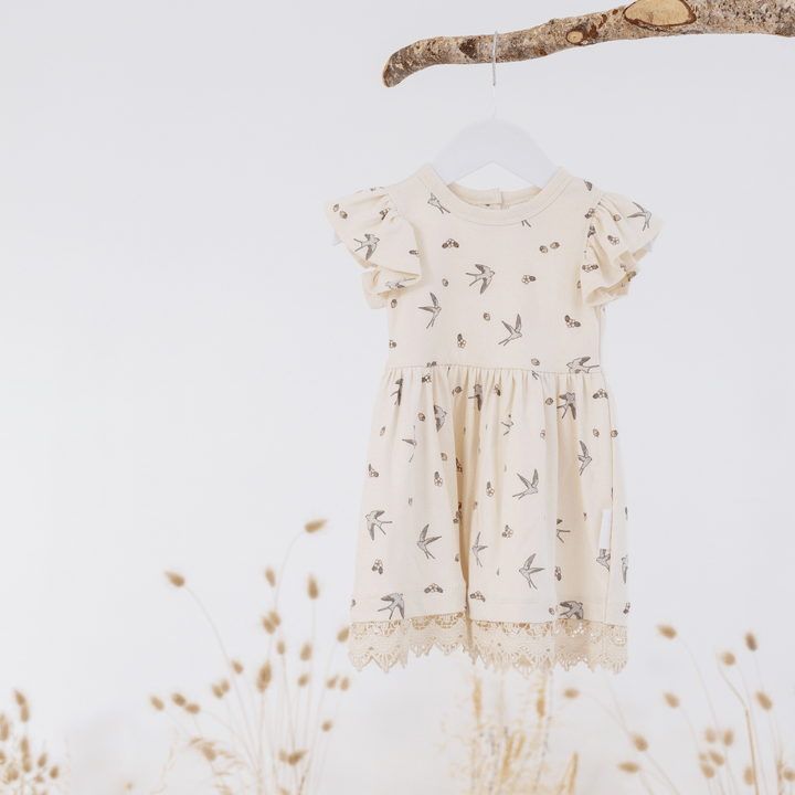 Off-white swallow and strawberry-print dress with lace trim and ruffle sleeves, on a hanger, hanging from a tree branch, with dried flowers below