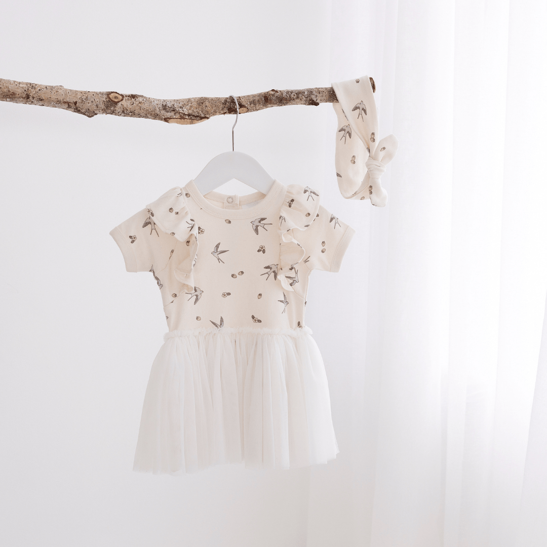 Beautiful dress for babies and kids featuring a delicate hand-drawn swallow and strawberry print on the top half, short sleeves with a ruffle at the shoulder, and a tulle skirt, hanging on a hanger from a branch, with a matching headband beside it