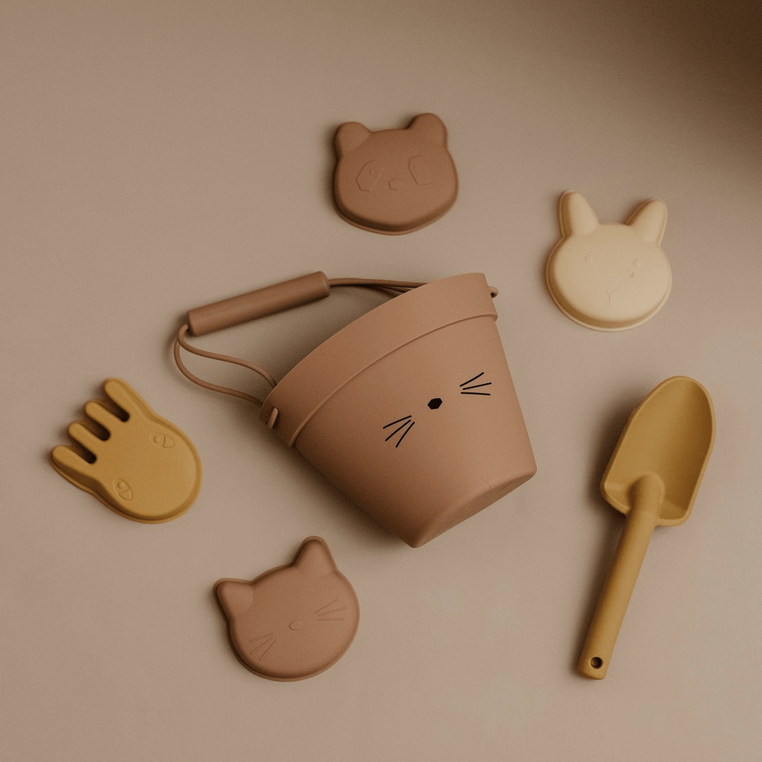 An eco-friendly tan bucket containing beach toys including the Classical Child Silicone Sand Set, shovels, and cat toys, arranged neatly on a table.