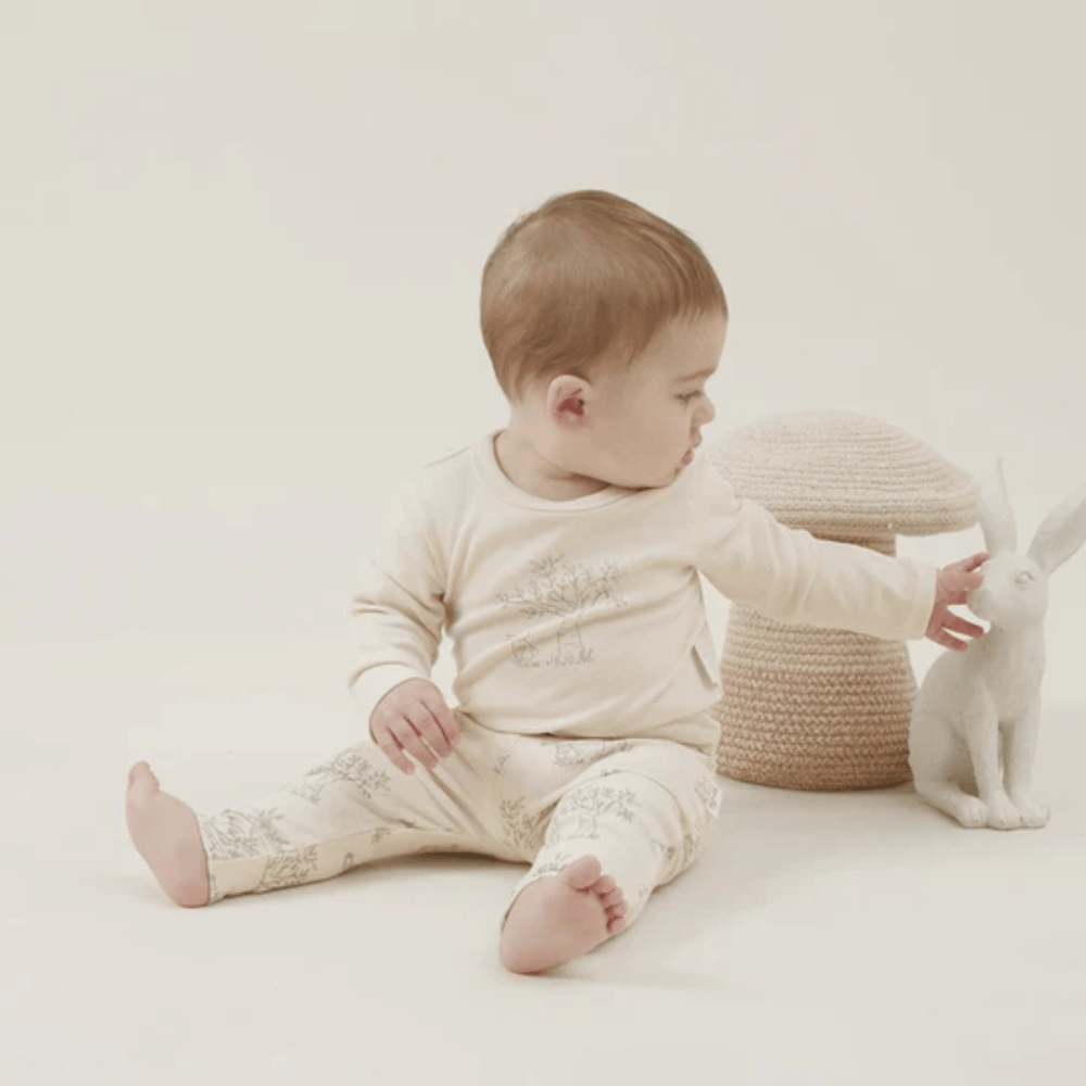 A baby, dressed in an Aster & Oak Organic Bunny Luxe Rib Long-Sleeved Top, sitting and reaching out towards a toy rabbit on a neutral background.